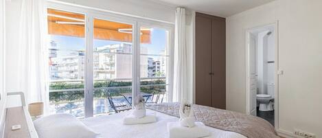 A hotel room with a bed, two white towels, a window with a view of buildings, and a door leading to a bathroom.