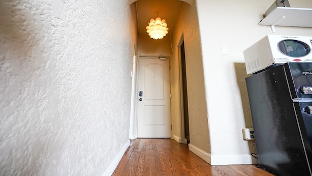 Un couloir étroit, avec une porte blanche au fond, un réfrigérateur noir doté d’un compartiment congélateur en haut, et un luminaire fixé au plafond.
