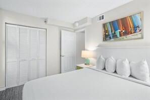 Ein Hotelzimmer mit einem Bett, weißen Kissen, einem Nachttisch mit einer Lampe, einer Tür und einem gerahmten Bild an der Wand.