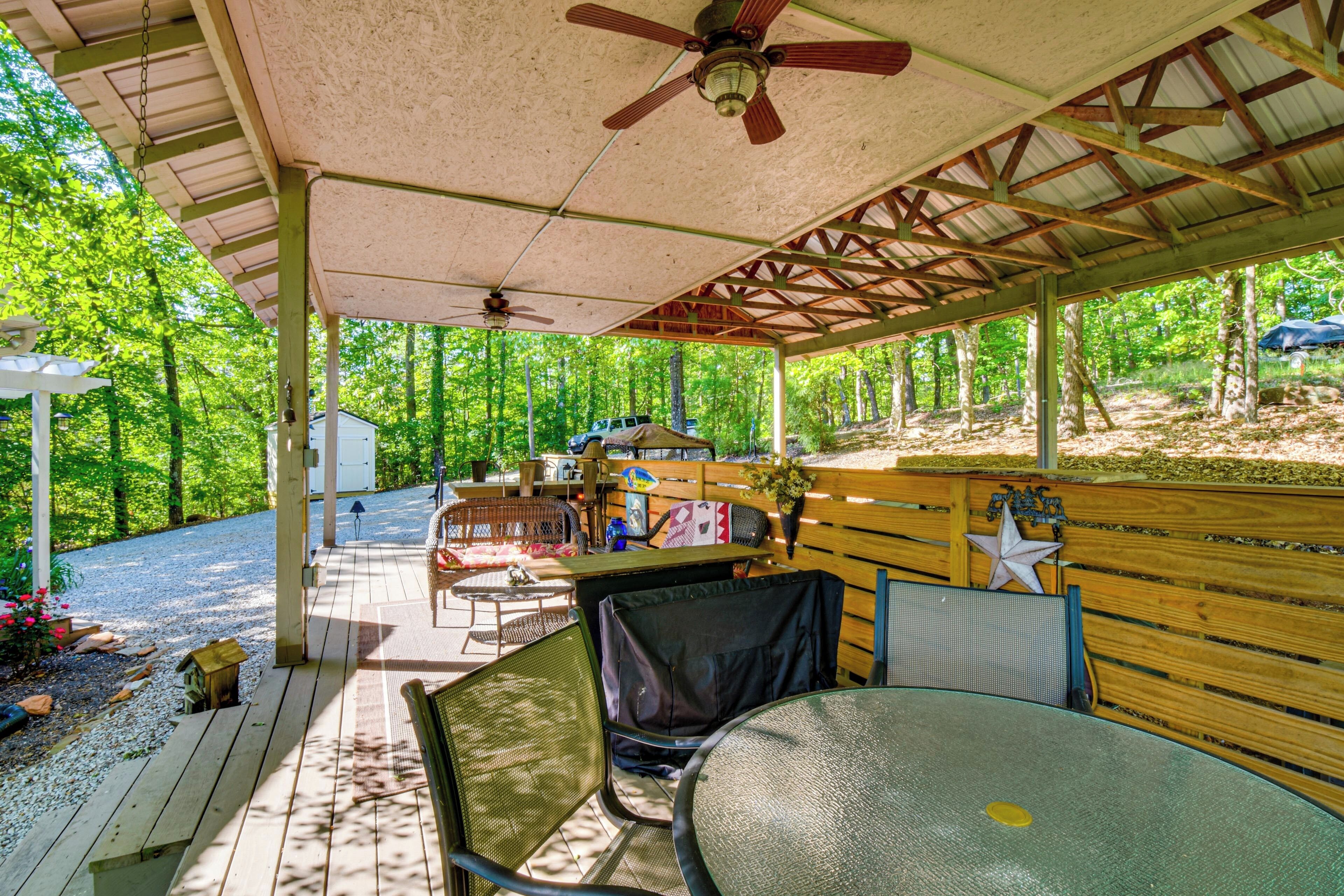 A covered outdoor area with a table and chairs, a ceiling fan, and a view of a forested area.