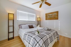 Une chambre à coucher avec un lit, un ventilateur de plafond, une fenêtre avec des stores, une table de chevet avec une lampe et un tableau au mur.