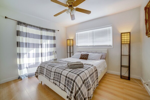 Un dormitorio con cama, ventilador de techo, ventana con persianas y lámparas de noche.