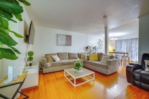 A living room with a sectional sofa, a glass coffee table, a TV mounted on the wall, and a dining area in the background. - Family Gem w/ Pet-Friendly Yard! 8 Mi to Riverside (Moreno Valley)