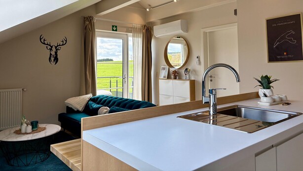 Une chambre d’hôtel moderne dotée d’une lucarne, d’une grande fenêtre donnant sur un champ verdoyant, d’un miroir rond, d’un lavabo avec un robinet contemporain et d’un lit avec une literie de couleur turquoise.