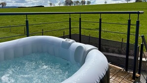 Un bain à remous extérieur doté d’une rambarde en métal, entouré d’un champ herbeux et d’arbres, sous un ciel nuageux.