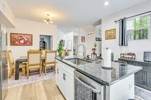 Una cocina moderna con una isla central, un comedor con sillas de madera y un amplio ventanal con vistas a árboles.
