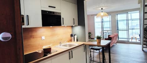 A modern kitchen with white cabinets, a wooden countertop, and a sink. There is a dining area with a table and chairs, and a large window providing natural light.