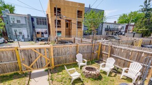 Une cour arrière clôturée, avec des chaises en plastique blanches, un foyer et une terrasse en bois.