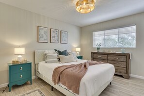 Ein Schlafzimmer mit Bett, Nachttischen, einem Schminktisch, Wandbildern und einem Fenster mit Jalousien.