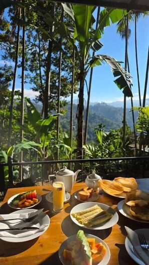 A table with breakfast items, a teapot, and a glass of juice, set against a backdrop of tall trees and a mountainous landscape. - Mango shadow inn  (Ella)