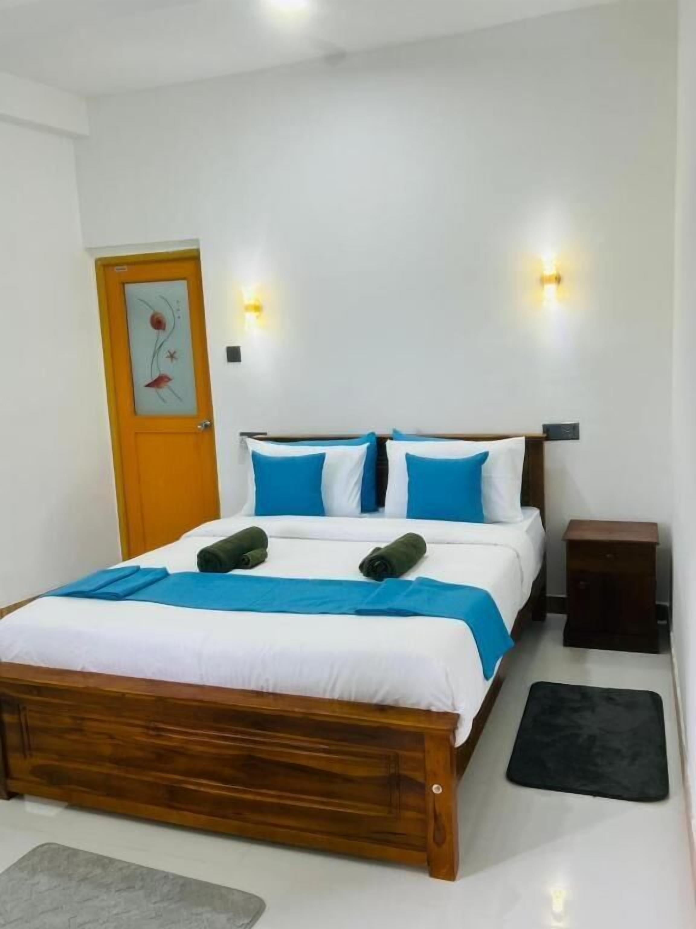 Une chambre avec un lit en bois, du linge de lit blanc, des coussins bleus, deux lampes fixées au mur, une table de chevet en bois et un tapis noir.