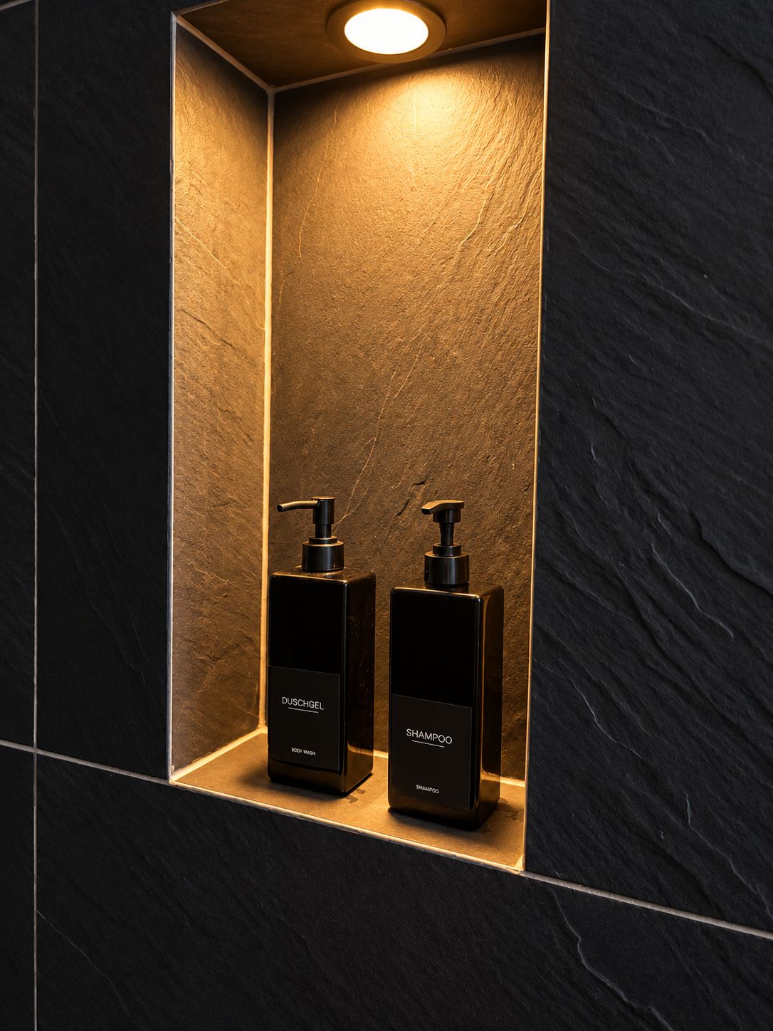 Une salle de bain avec un mur recouvert de carreaux sombres et une étagère sur laquelle reposent deux flacons noirs de gel douche et de shampoing de la marque Duschgel.