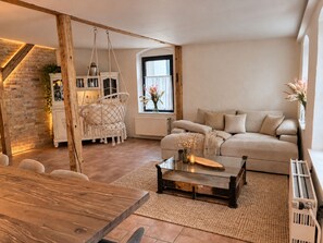 A cozy living room with a wooden table, a hanging chair, a sofa, and a window with flowers.