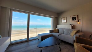 Un salon moderne avec une grande porte-fenêtre coulissante donnant sur la plage, un canapé gris, une petite table et un lampadaire.