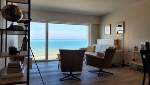 Un salon moderne avec une grande fenêtre donnant sur la plage, un globe terrestre posé sur une étagère et un bureau avec des livres.