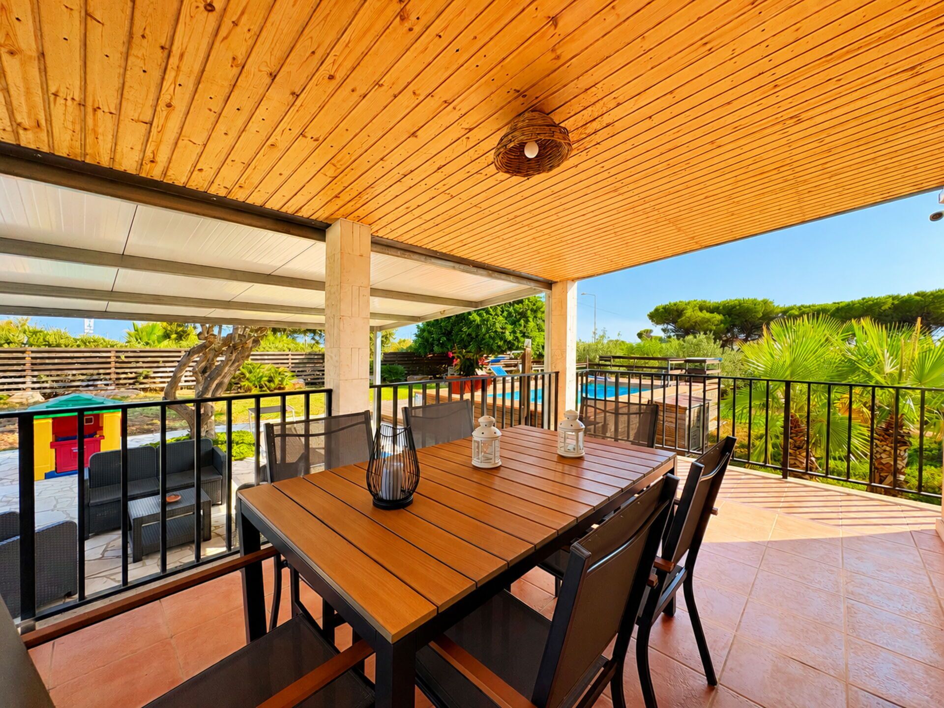 An outdoor dining area with a wooden table and chairs, a view of a swimming pool, and a garden with various plants.