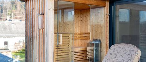 A wooden sauna with glass doors, a bench inside, and a chair outside.