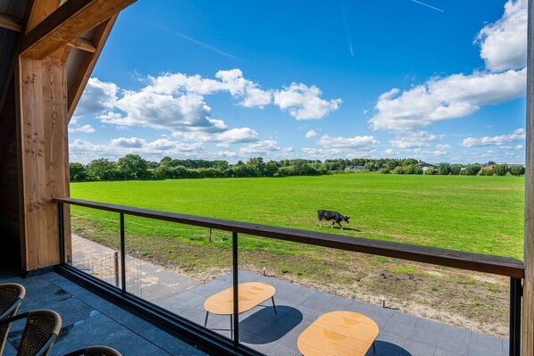 A balcony with a view of a green field and a cow, wooden furniture, and a clear sky.