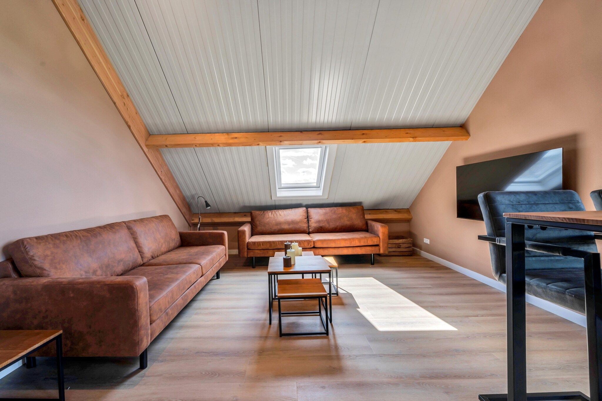 Een loft-achtige woonkamer met een hoge plafond, houten balken, een groot raam, een flatscreen-tv, leren banken en een salontafel.