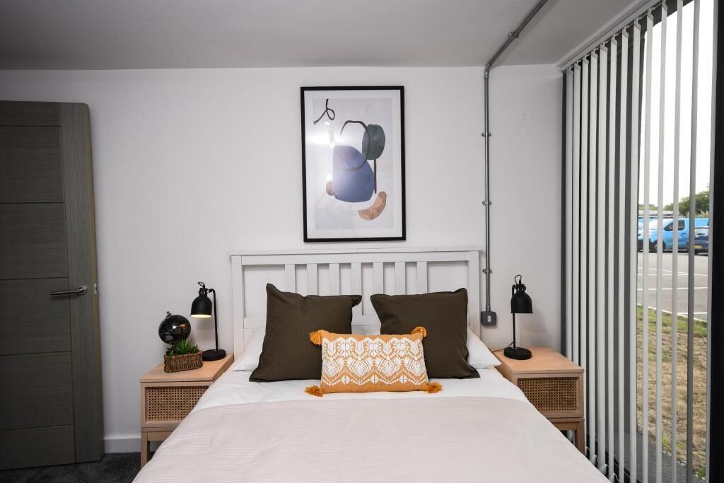 A bedroom with a white bed, two brown pillows, a patterned pillow, two bedside tables with lamps, a framed picture on the wall, and a view of the street through the window.
