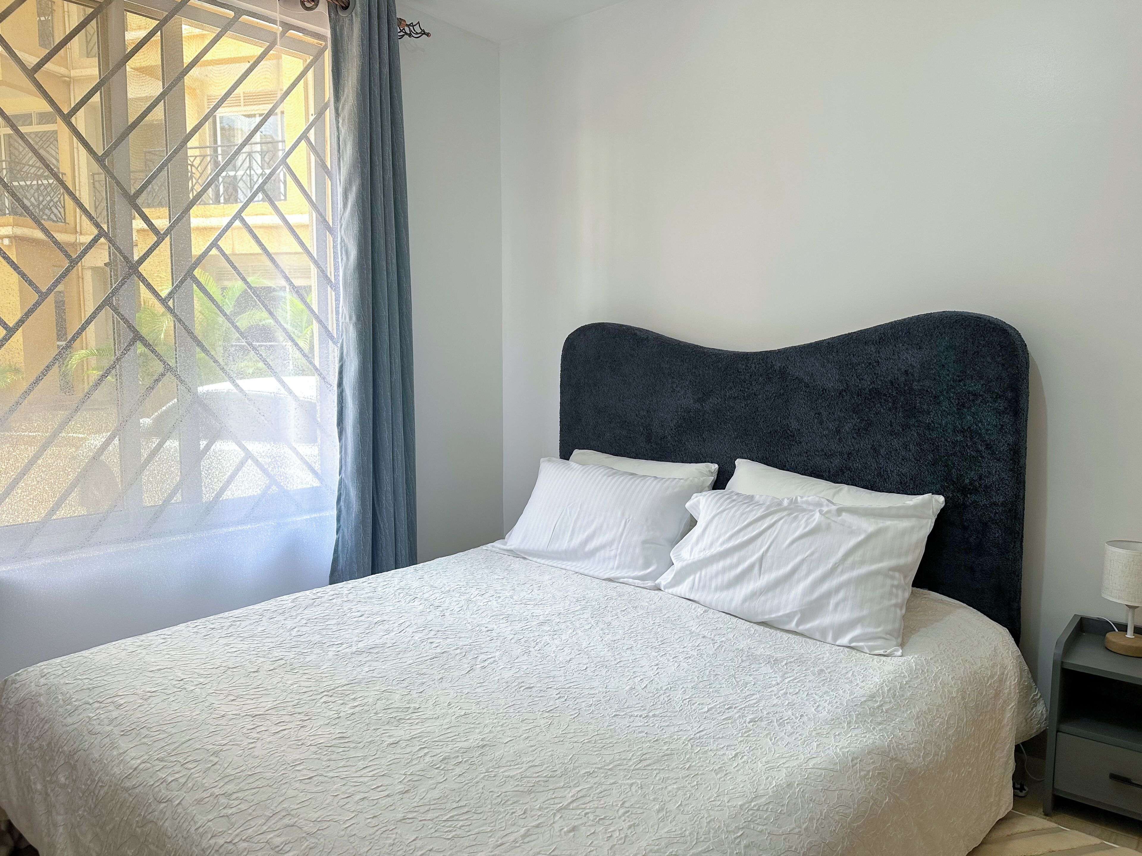 A neatly made bed with a textured white bedspread and two pillows, a dark headboard, a bedside table with a lamp, and a window with a curtain.