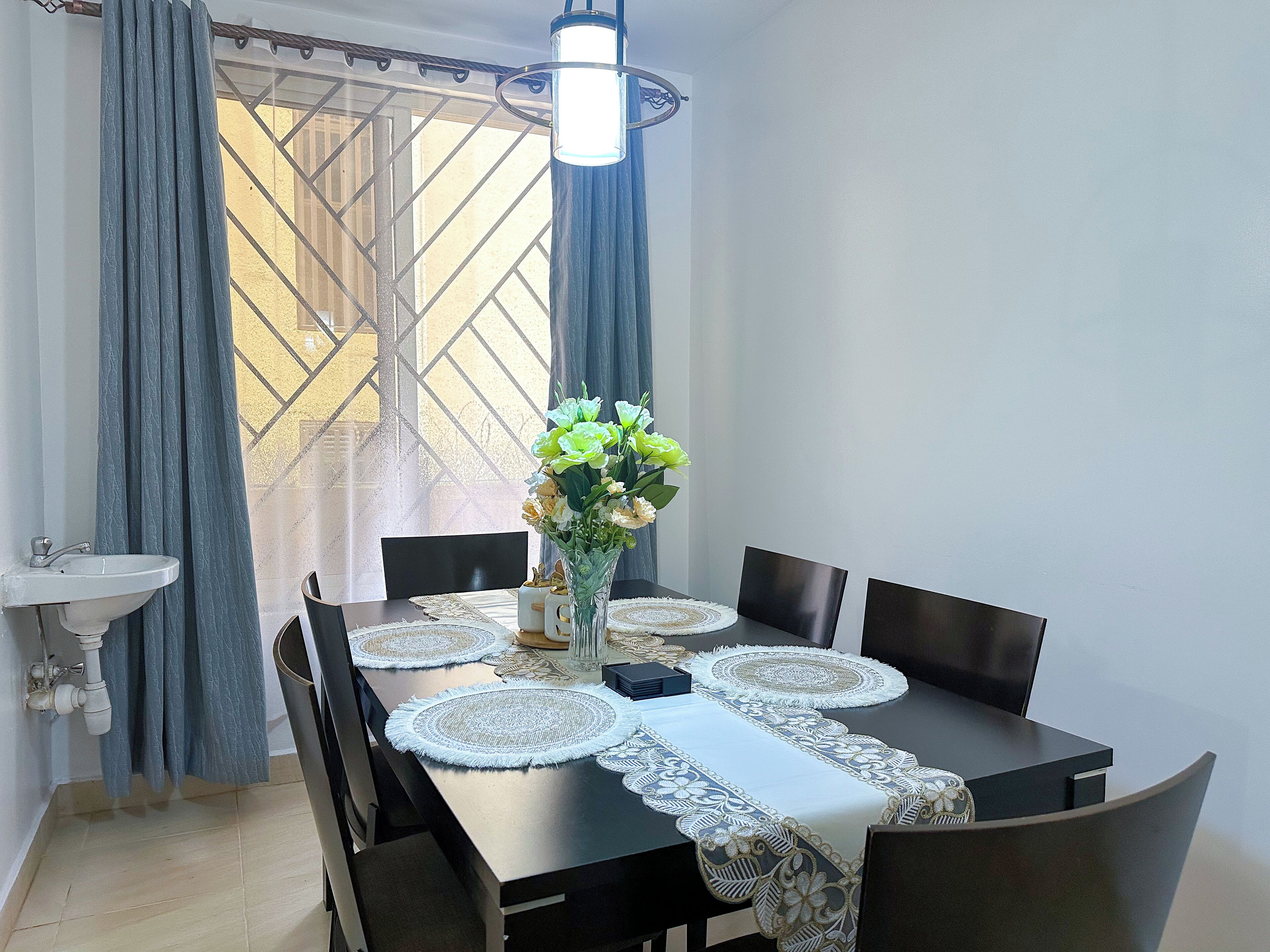 A dining table with a white tablecloth, surrounded by chairs, a vase with flowers, and a pendant light.