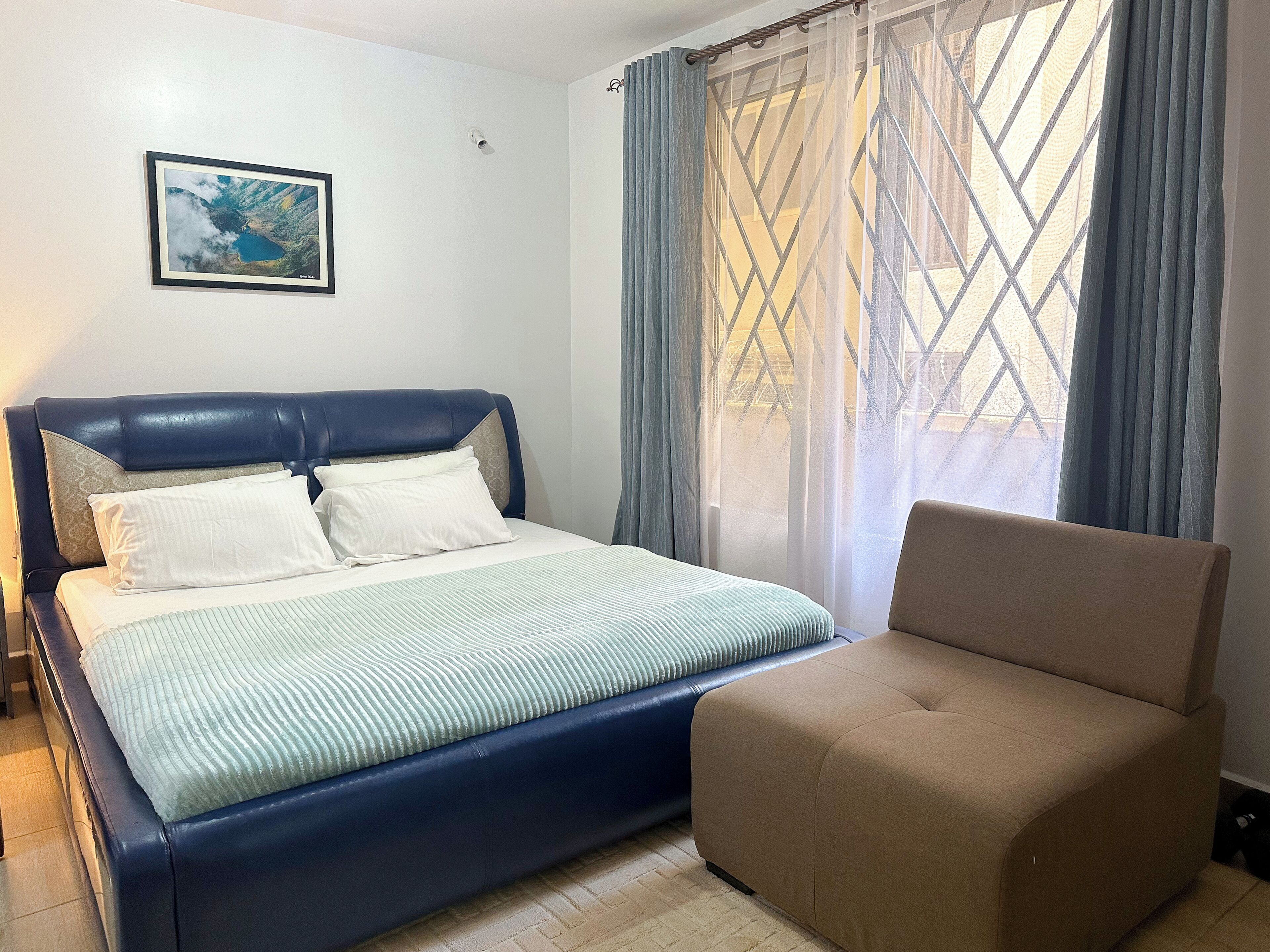 A bedroom with a bed, a chair, a window with curtains, and a framed picture on the wall.