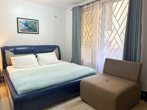 Ein Schlafzimmer mit einem Bett, einem Sessel, einem Fenster mit Vorhängen und einem gerahmten Bild an der Wand.