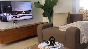 A living room with a flat-screen TV, a brown sofa, a coffee table with books and a decorative item, a wooden sideboard, and a large green plant. - The snug cove, beautifully furnished and fully functional. Family friendly! (Najjera)