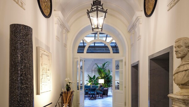 A hallway with a marble column, a bust sculpture, a table with chairs, and a view into a dining area with tables and chairs.