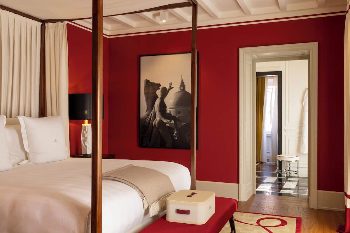 A hotel room with a four-poster bed, a red wall, a framed artwork, and a small bedside table with a lamp.