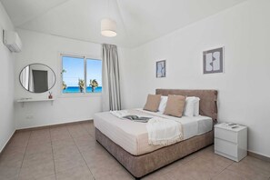 A bedroom with a bed, a bedside table, a window with a view of palm trees, a mirror, and a ceiling light. - Aria Del Mar Villa by Nomads (Paphos)