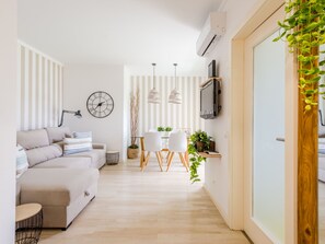 Ein modernes Wohnzimmer mit Sofa, Essbereich mit Stühlen, einer Uhr, Pendelleuchten und einem an der Wand montierten Fernseher.