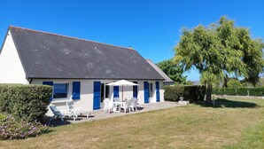 Une maison avec un toit en pente, une terrasse avec des meubles d’extérieur et un grand arbre en arrière-plan.
