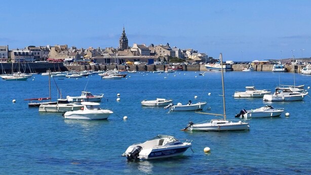 Un port où de nombreux bateaux sont amarrés, une église dont le clocher se détache dans le paysage, et une ville avec des bâtiments qui bordent le front de mer.