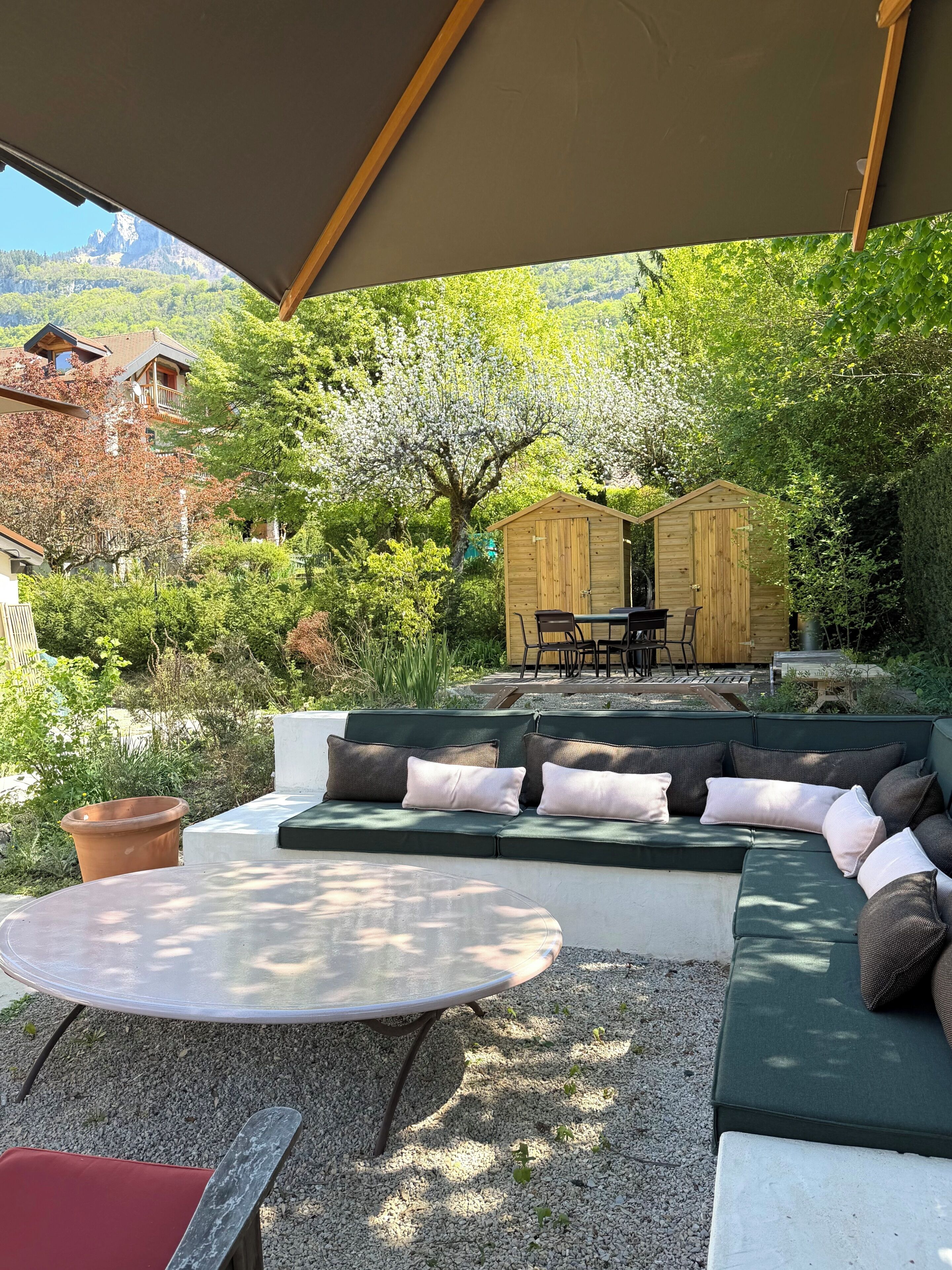 A covered outdoor seating area with a table, a sofa, and a wooden shed in a lush garden setting.