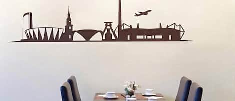 Un comedor con una mesa preparada para dos personas, sillas y un adhesivo mural que representa el horizonte de una ciudad.
