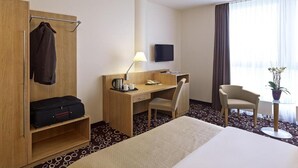 Et hotelværelse med en seng, et skrivebord med stol, et klædeskab, et fjernsyn og et lille bord med en plante.