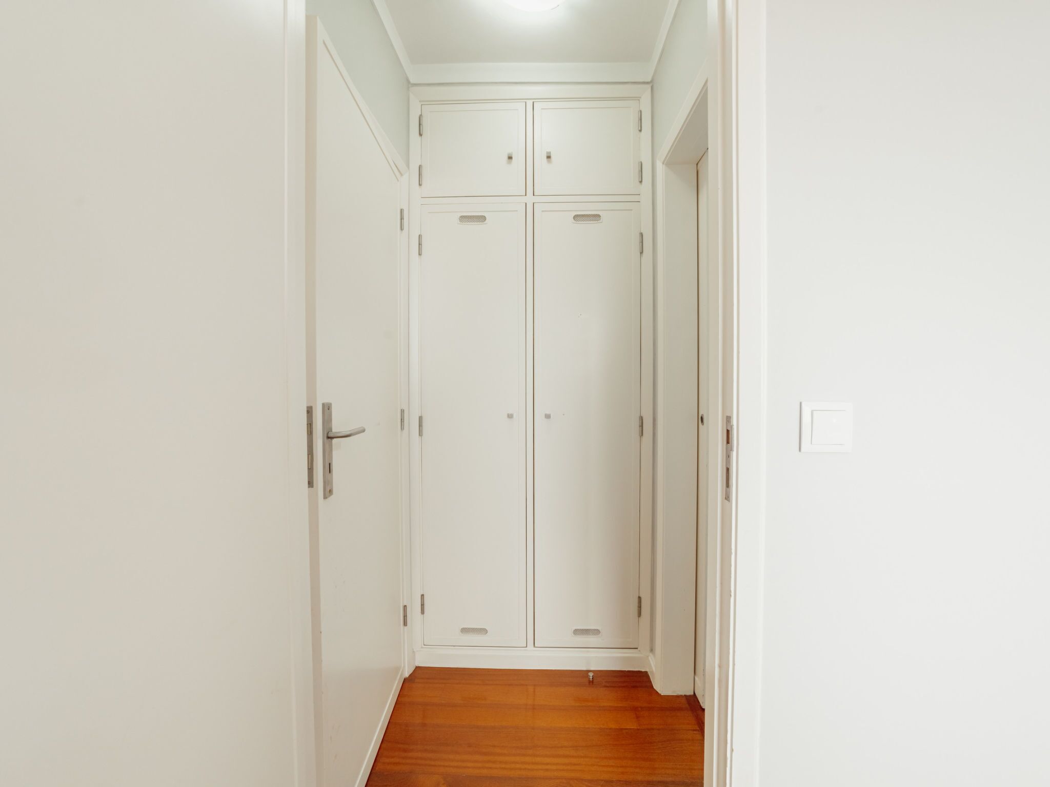 Un pasillo que conduce a una puerta doble blanca con pomo, un interruptor de luz y suelo de madera.