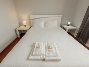 Habitación de hotel con una cama grande, dos mesitas de noche con lámparas y una toalla doblada con artículos de aseo sobre la cama.