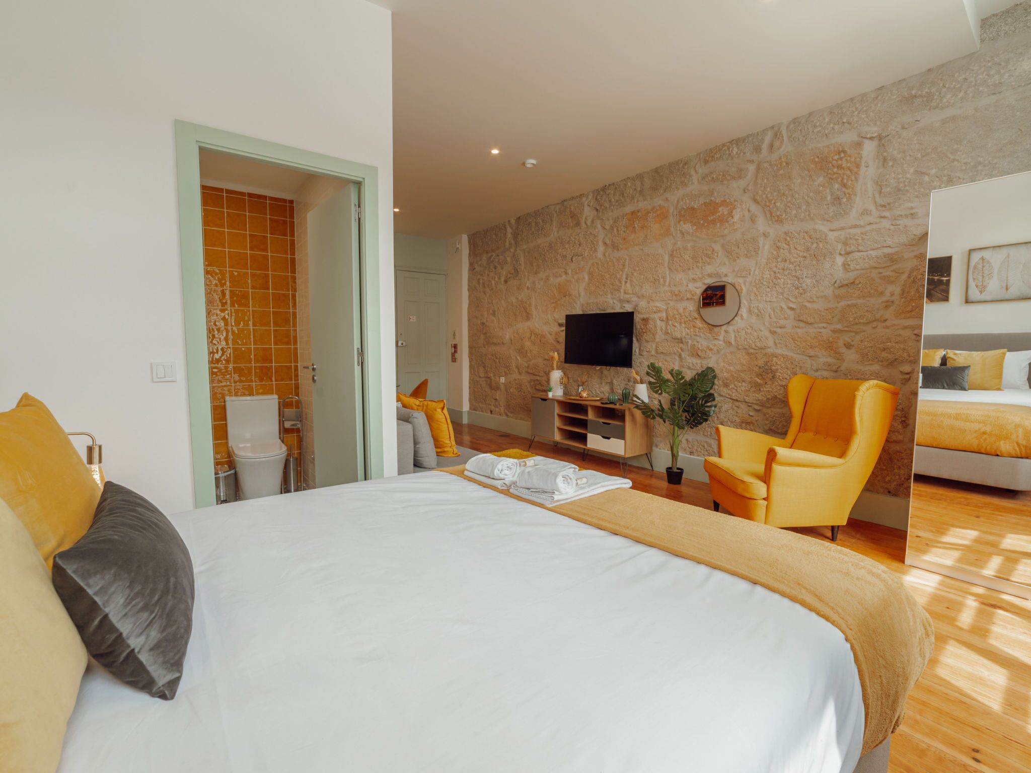 A hotel room with a large bed, a stone wall, a television, and a yellow armchair.