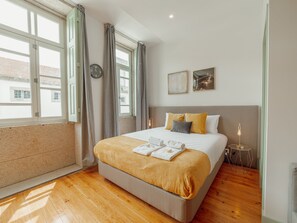 A hotel room with a large bed, a bedside table with a lamp, a window with curtains, and a view of buildings outside. - Apartment  by Interhome (Porto)