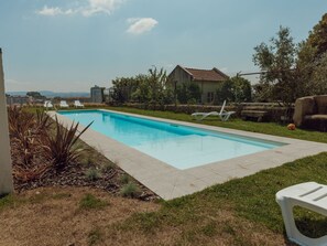 A rectangular swimming pool with a clear blue water surface, surrounded by a concrete deck. There are white lounge chairs and a concrete bench near the pool. The pool is set in a grassy area with a fence enclosing the property. - Apartment  by Interhome (Porto)