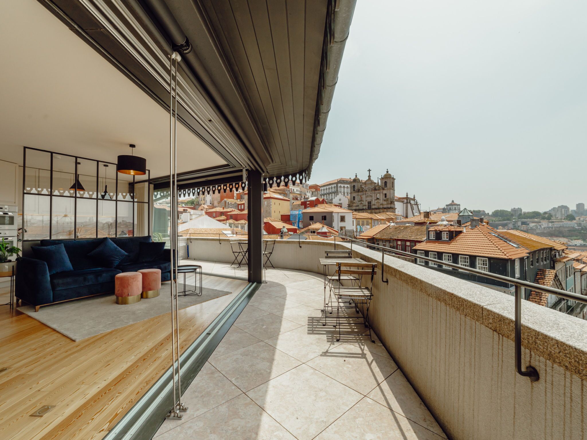 A modern balcony with a sofa and ottomans, overlooking a cityscape with historic buildings.