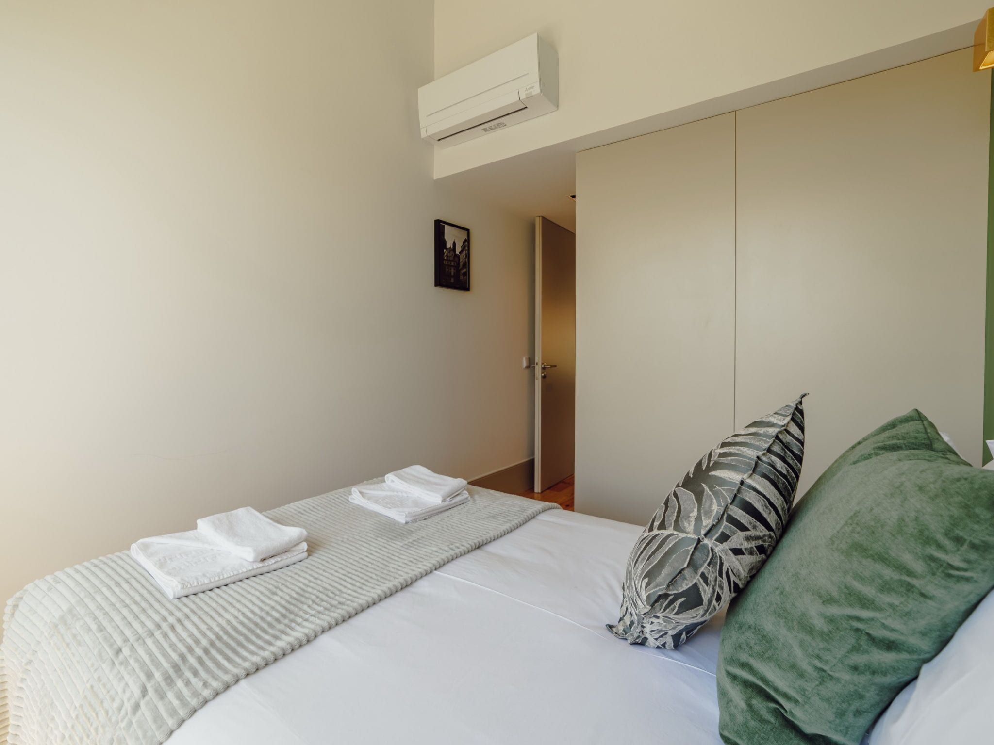 A bedroom with a bed, pillows, a blanket, a wall-mounted air conditioner, and a door leading to another room.