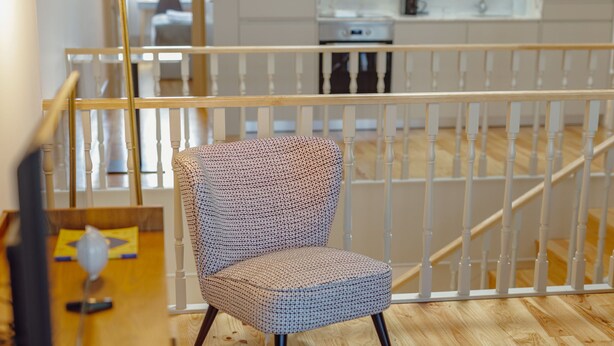 A chair with a patterned fabric, a wooden table with a lamp, and a white railing in a room with wooden flooring. - Apartment  by Interhome (Porto)