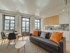 Ein modernes Wohnzimmer mit einem grauen Sofa, orangefarbenen Kissen, einem kleinen runden Tisch und Stühlen.
