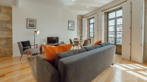 Een moderne woonkamer met een grijze bank, houten vloeren, een stenen muur en grote ramen.