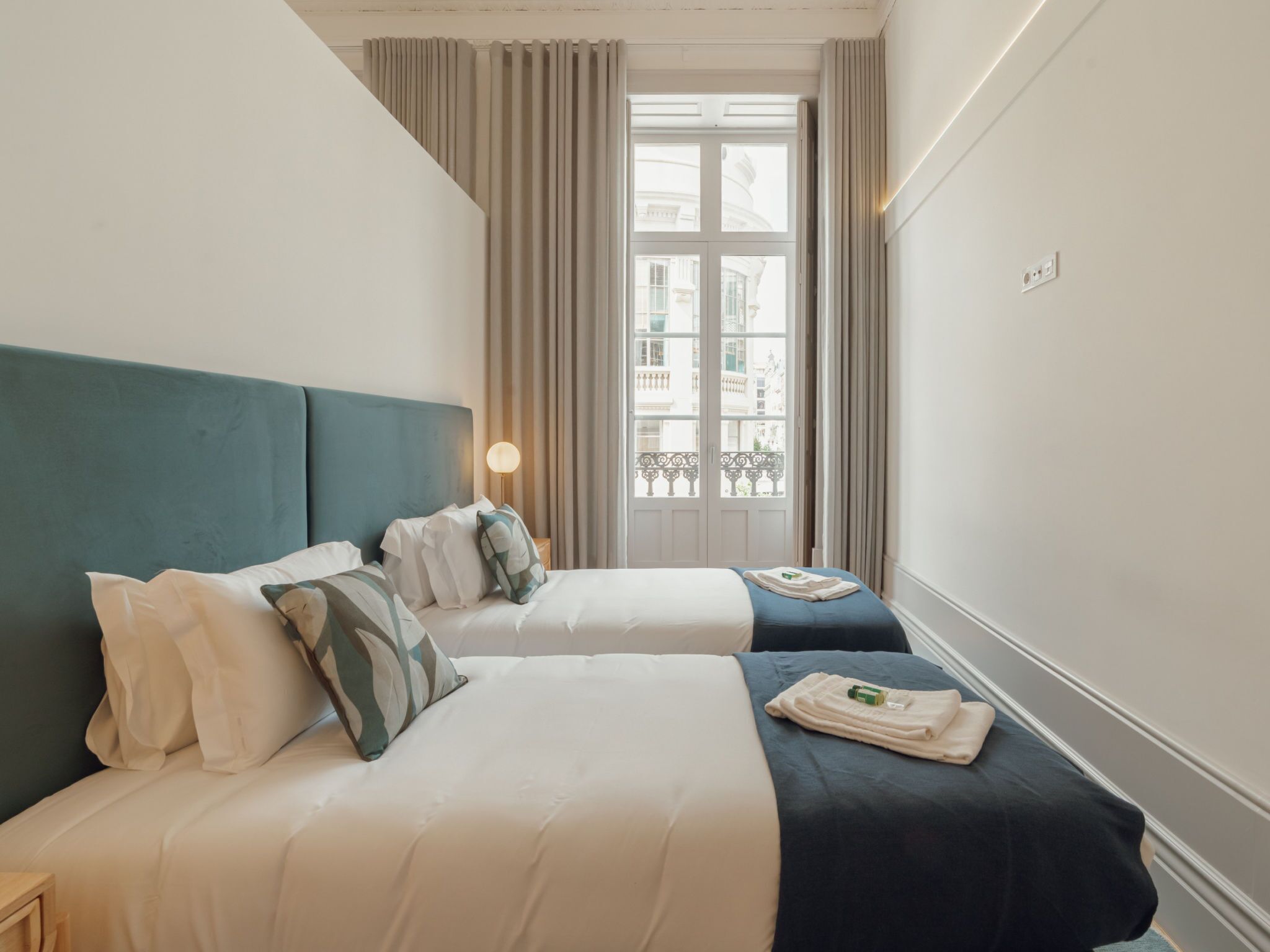 A hotel room with two beds, a window with curtains, a lamp, and a view of buildings outside.