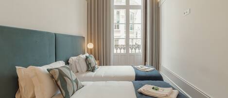 A hotel room with two beds, a window with curtains, a lamp, and a view of buildings outside.